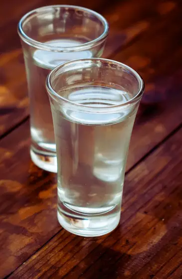 Two clear shot glasses filled with Chhaangster Raksi, served on a rustic wooden table.
